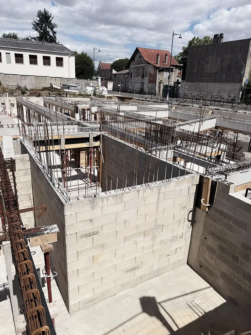 Chantier d’immeuble vu d’en haut : murs en blocs béton, poutres coffrées en bois, étais métalliques rouges, maisons voisines en arrière-plan.