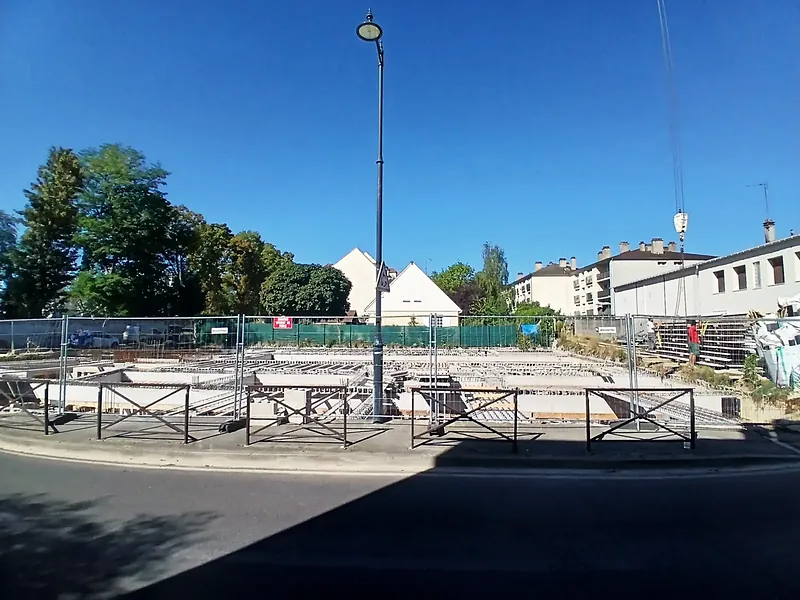Chantier palissadé vu depuis la rue, ciel bleu, maisons et immeubles voisins.