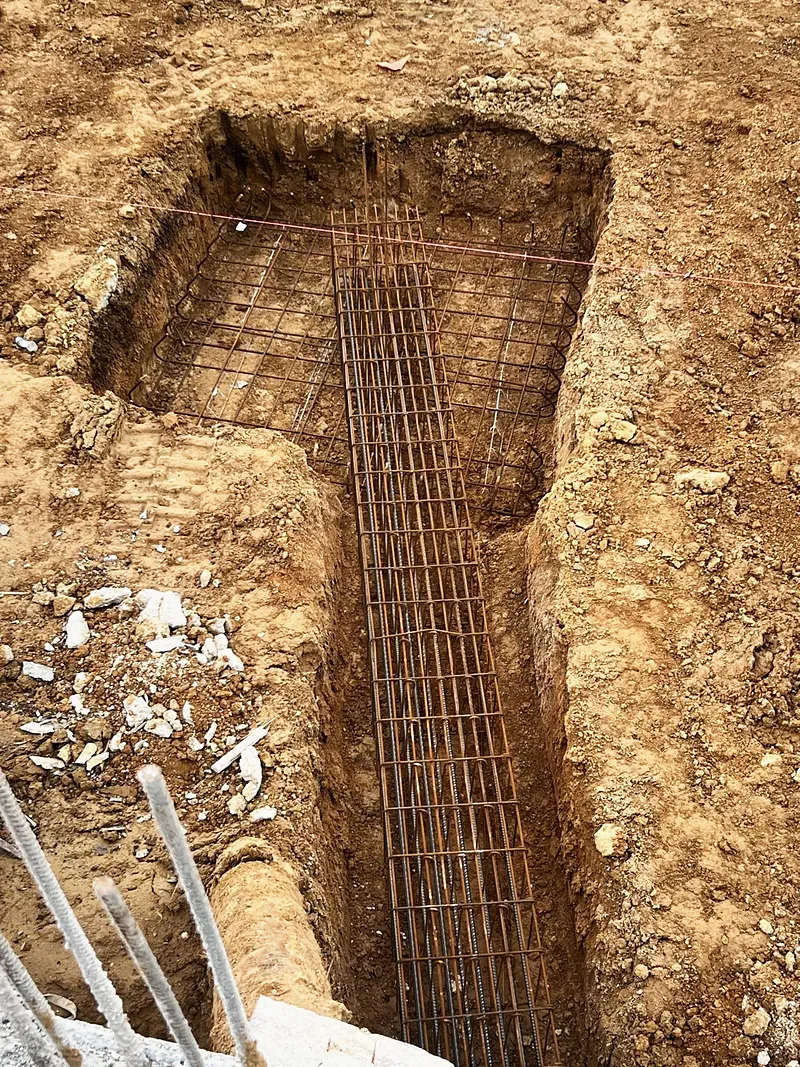 Tranchée de fondation avec armatures filantes reliées à une semelle carrée ferraillée.
