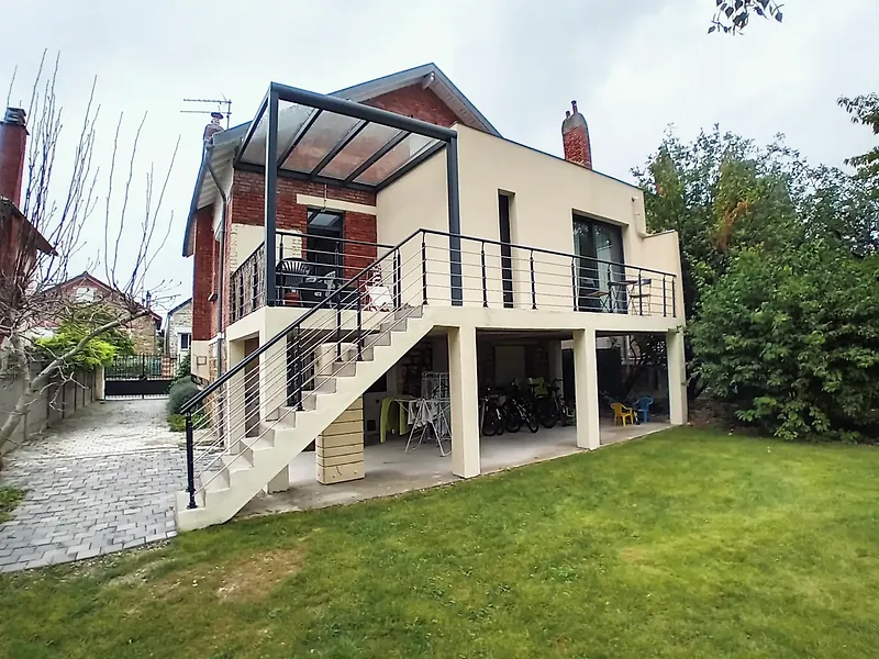 Vue de trois quarts de l’extension enduite beige, escalier en béton à garde-corps anthracite, pergola à couverture transparente, allée pavée et pelouse.