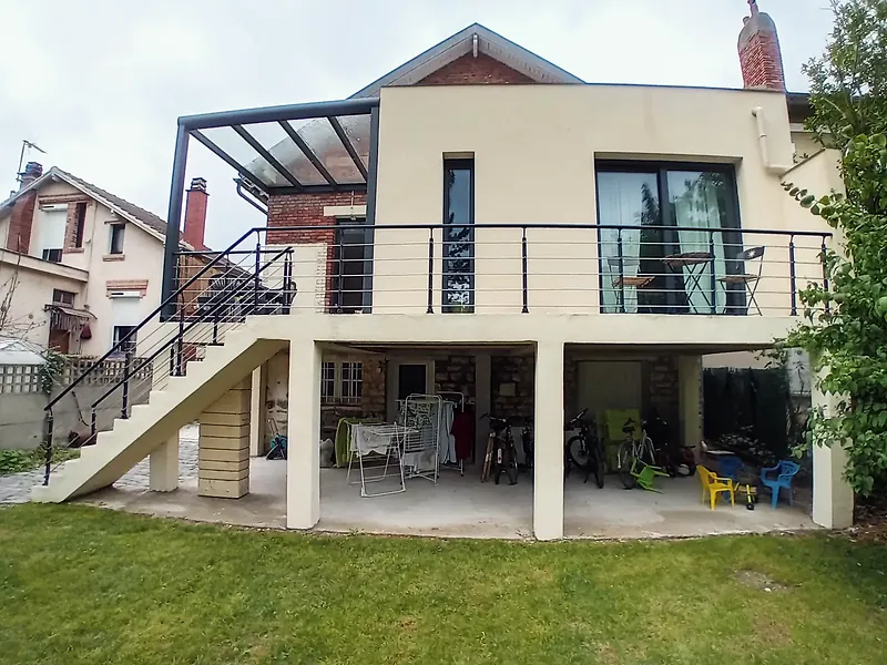 Façade arrière avec terrasse portée par des poteaux enduits, baie coulissante sombre et fenêtre verticale étroite, espace couvert sous la terrasse, pelouse au premier plan.
