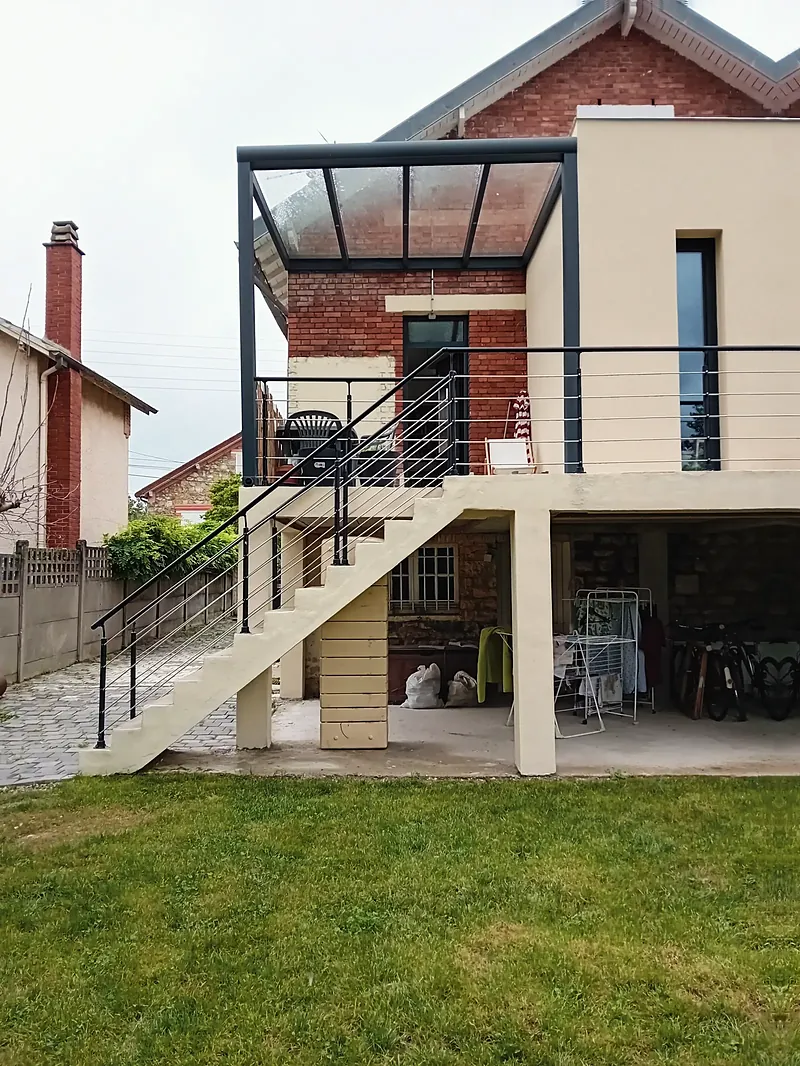 Pergola à poteaux et poutres anthracite et couverture transparente devant un mur de brique rouge, volume enduit beige à droite, escalier en béton au premier plan.