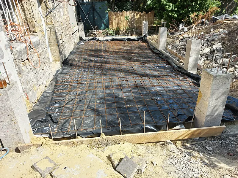 Treillis soudé posé sur un film plastique noir au pied d’un mur en moellons, poteaux en blocs béton avec fers en attente, rive coffrée en planche.