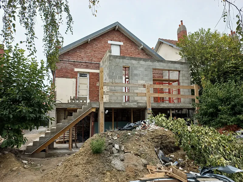 Volume en blocs béton monté contre une maison en brique, terrasse sur étais bordée de garde-corps provisoires en planches, escalier en béton sur coffrage bois, terre et gravats au premier plan.