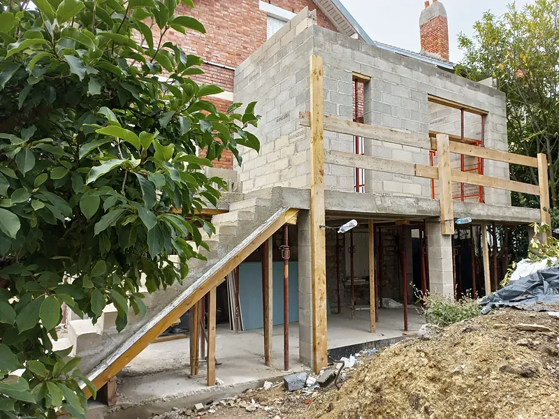 Vue de biais du volume en blocs béton, dessous de la terrasse soutenu par des étais métalliques et des poteaux en blocs, escalier en béton coffré.
