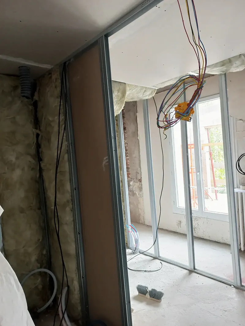 Pièce en travaux avec montants métalliques de cloison, laine minérale contre le mur, gaines électriques en attente et porte-fenêtre blanche.