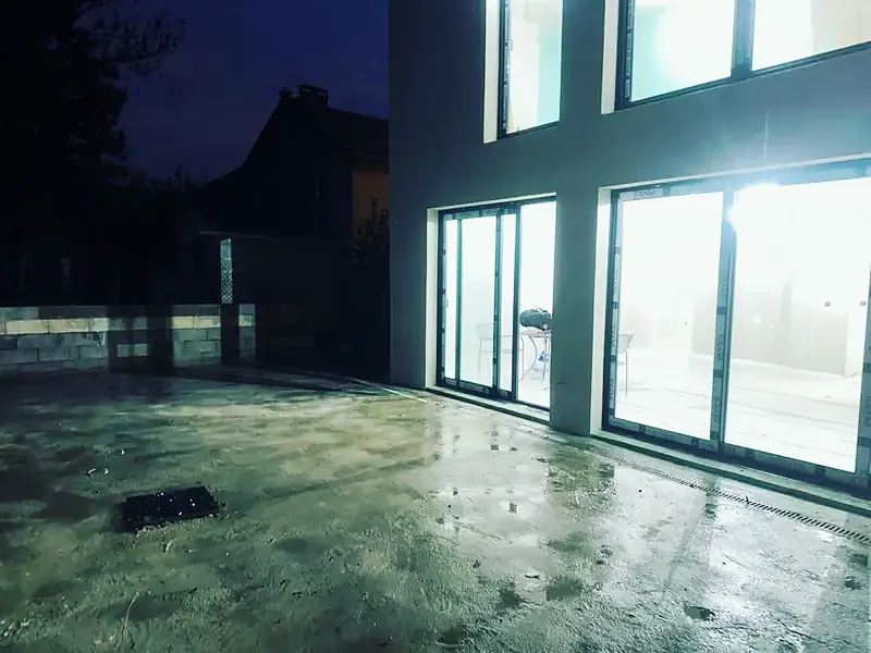 Terrasse en béton de nuit devant des baies vitrées éclairées.