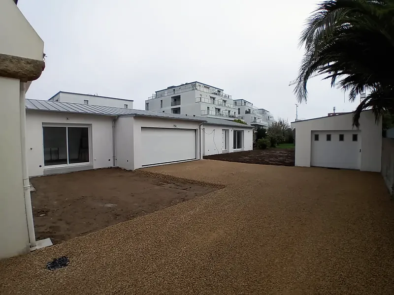 Cour gravillonnée bordée par l’extension blanche à porte de garage et par un garage indépendant en fond de parcelle, palmes au premier plan.