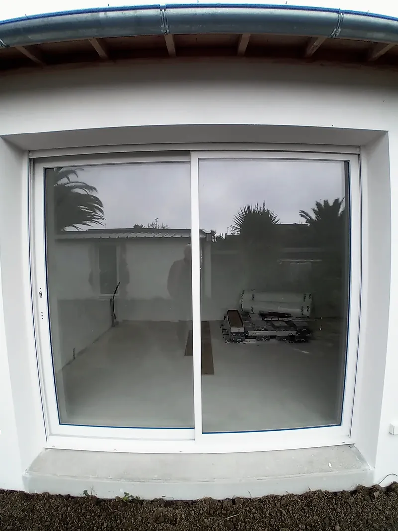 Baie coulissante blanche à deux vantaux dans une façade enduite blanche, seuil en béton, bord de toiture et gouttière au-dessus.