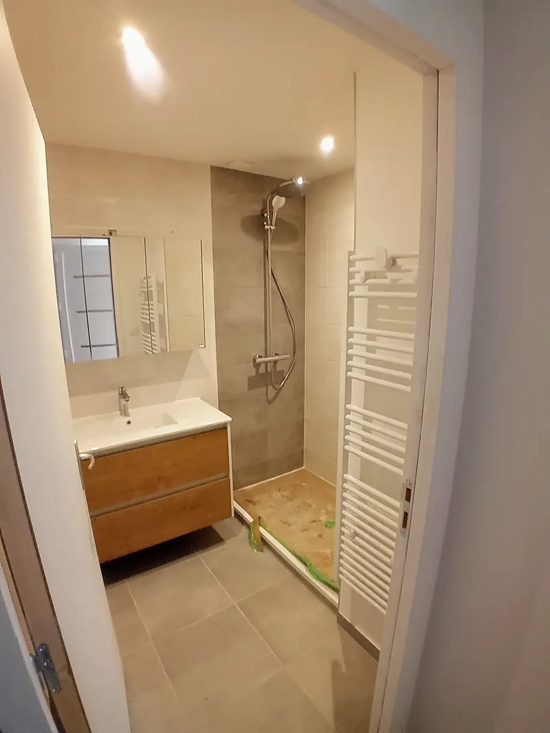 Salle d’eau carrelée gris clair avec douche à l’italienne, colonne de douche, meuble vasque en bois clair, miroir et sèche-serviettes blanc.
