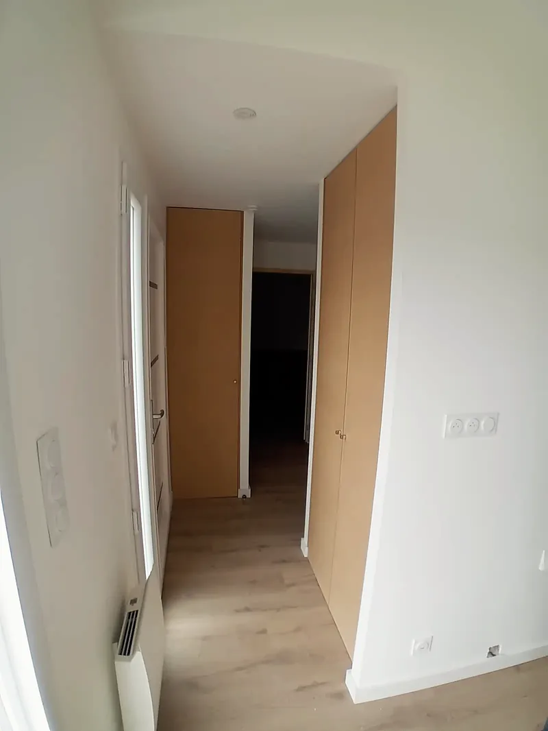 Couloir blanc au sol en lames de bois, placards à portes en bois clair, radiateur électrique et prises murales.