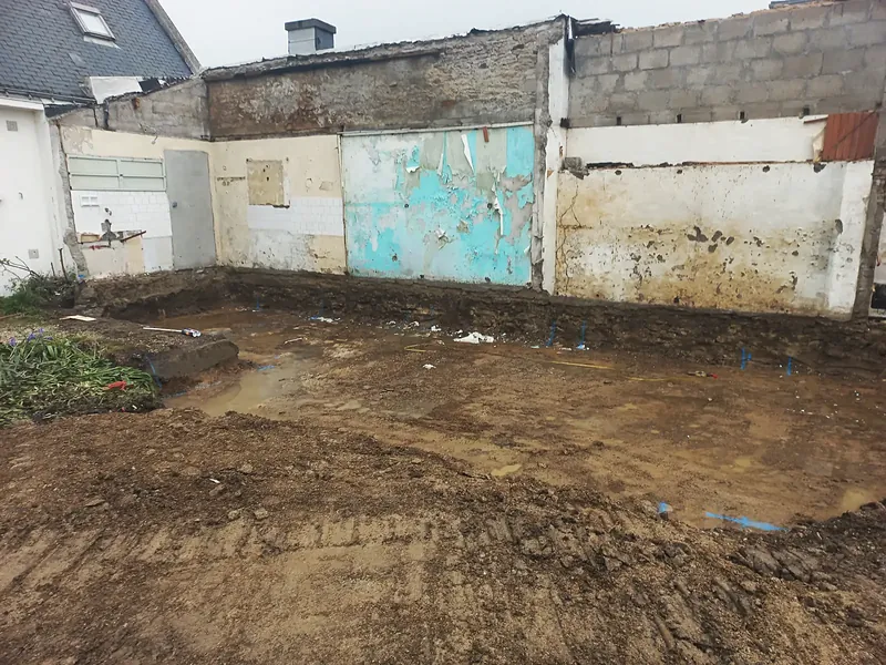 Terrain décapé et détrempé devant un mur ancien couvert de faïence blanche, de peinture turquoise écaillée et d’enduits jaunes.