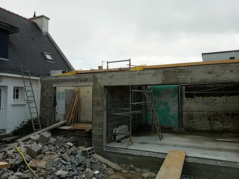 Murs en blocs béton avec une large ouverture surmontée d’une poutre en béton, échafaudage et échelles, gravats au premier plan.