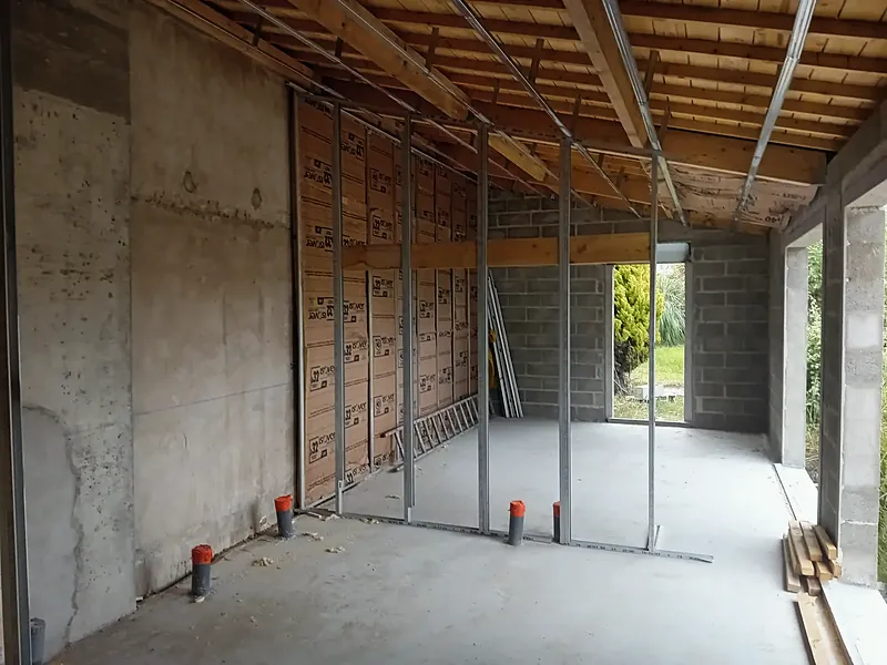 Pièce brute avec mur en béton, montants métalliques de cloison et rouleaux d’isolant en laine minérale, plafond à solives bois.