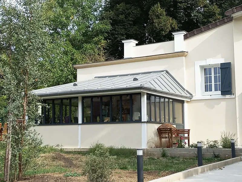 Extension vitrée de plain-pied aux menuiseries anthracite sur allège enduite crème, couverture métallique grise à joints debout, maison enduite à volets gris-bleu à droite, jardin planté au premier plan.