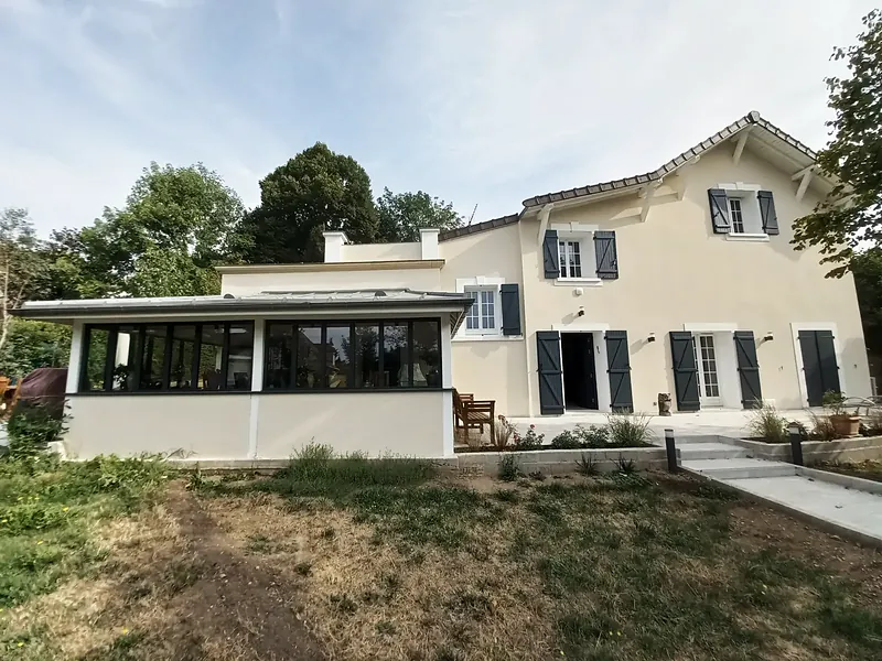 Façade sur jardin d’une maison enduite crème à volets gris-bleu, extension vitrée basse à gauche, terrasse et escalier, pelouse au premier plan.