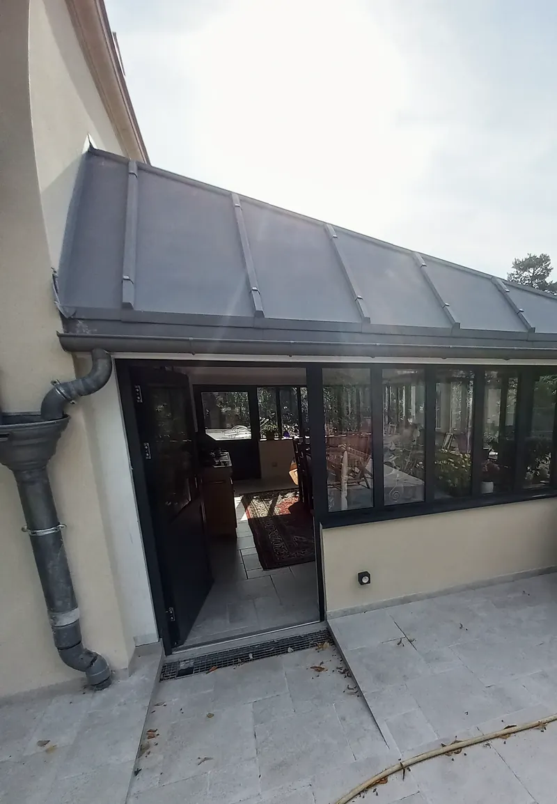 Angle de l’extension vitrée : couverture métallique grise à joints debout, gouttière et descente grises, porte ouverte sur la salle à manger, terrasse en dalles claires.
