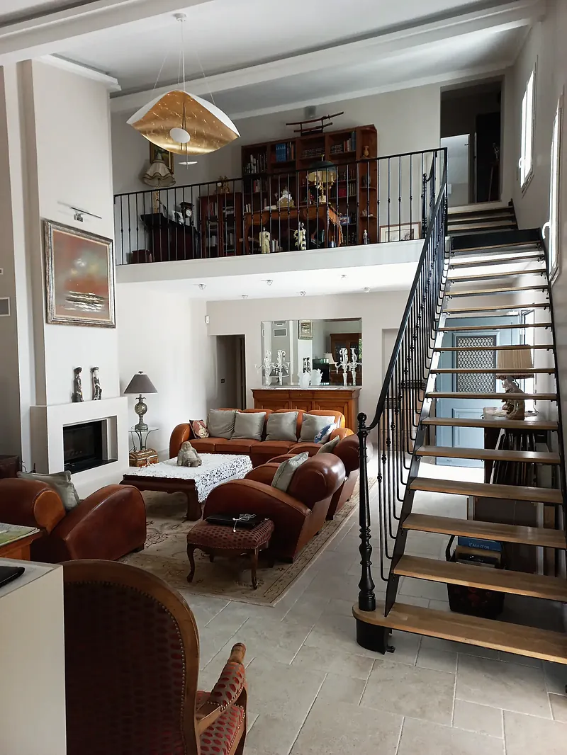 Séjour en double hauteur avec mezzanine bibliothèque bordée d’un garde-corps à barreaux noirs, escalier à limon noir et marches en bois, cheminée blanche, carrelage clair.
