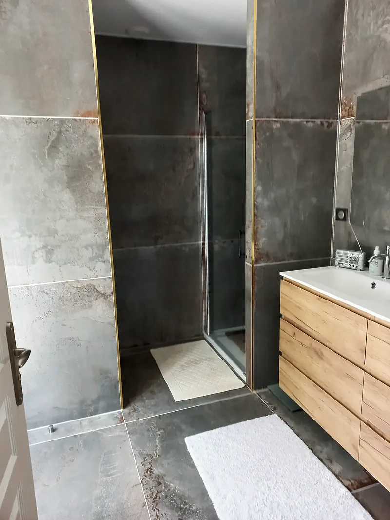 Salle d’eau au carrelage grand format gris veiné de rouille, profilés dorés, douche vitrée, meuble vasque en bois clair.