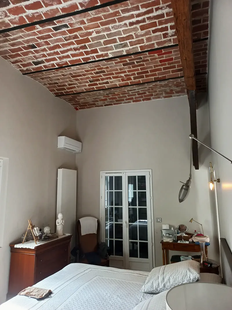 Chambre au plafond en voûtains de brique rouge entre poutrelles métalliques, poteau en bois sombre, porte-fenêtre blanche à petits bois.