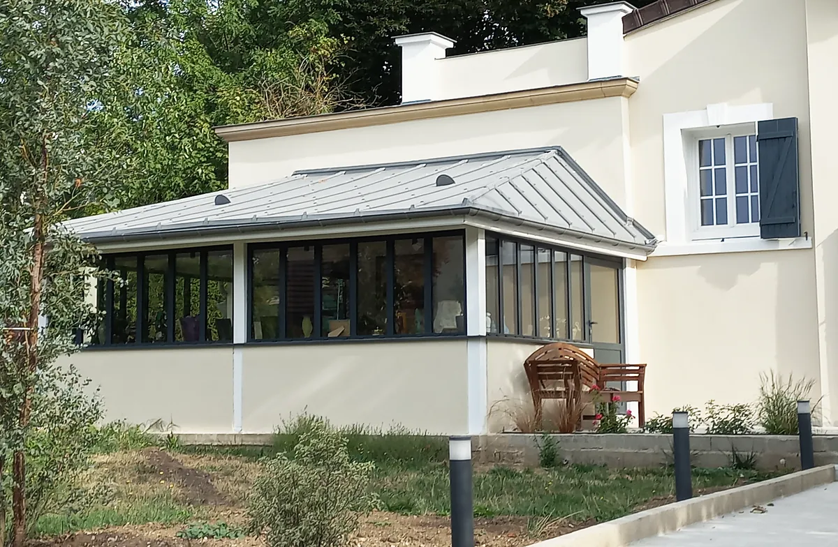 Extension vitrée de plain-pied aux menuiseries anthracite sur allège enduite crème, couverture métallique grise à joints debout, maison enduite à volets gris-bleu à droite, jardin planté au premier plan.
