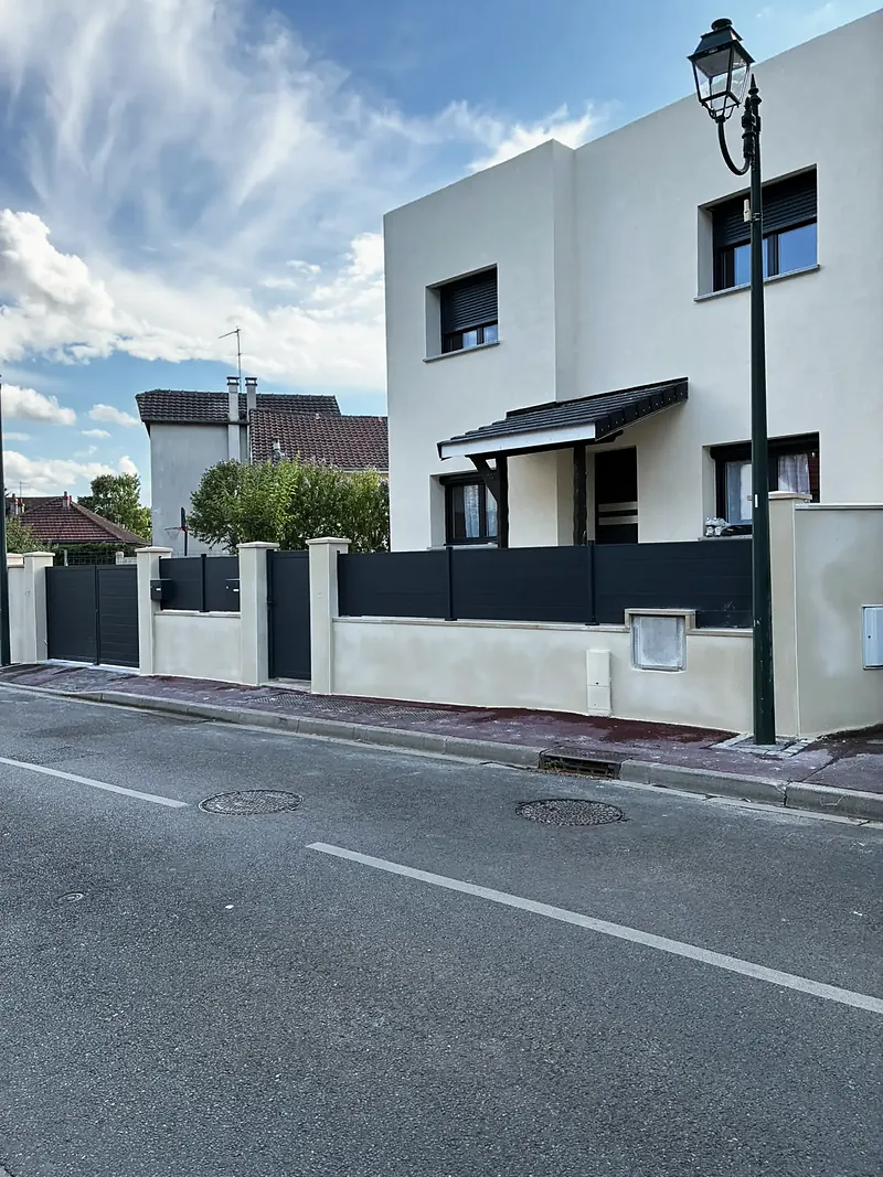 Façade sur rue de la maison achevée, auvent d’entrée et clôture à lames horizontales anthracite.