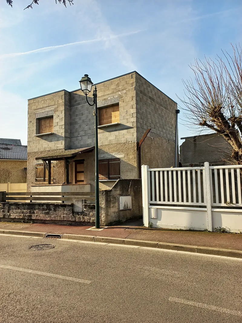 Maison cubique en parpaings bruts non enduits, fenêtres fermées par des panneaux de bois, clôture blanche.