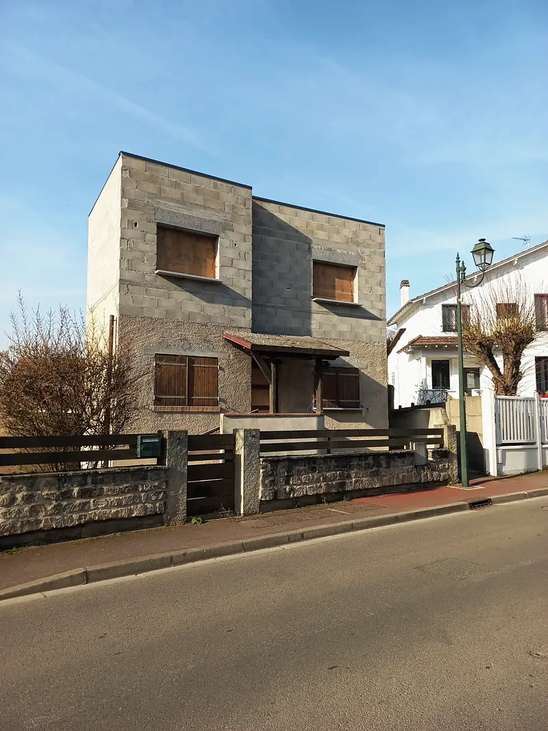Maison en blocs béton bruts vue de face, auvent en bois, muret de pierre et clôture bois.