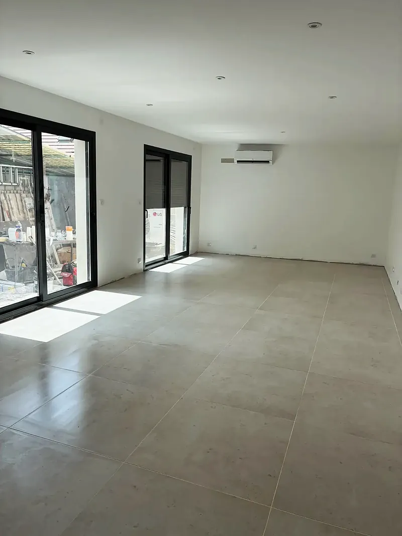 Grande pièce de vie vide et lumineuse, carrelage clair grand format, deux baies vitrées noires.