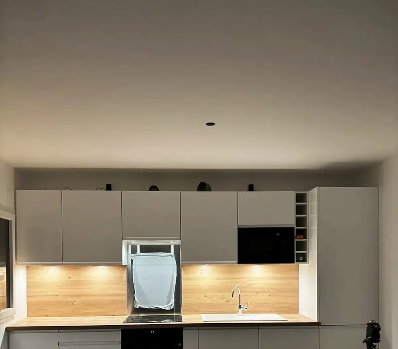 Cuisine blanche linéaire avec plan de travail et crédence en bois éclairés par des réglettes.