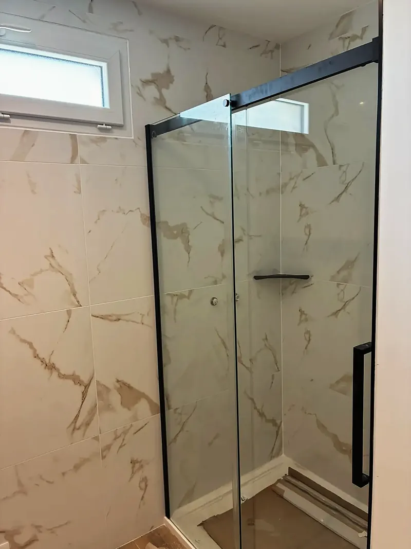 Douche avec paroi vitrée à profilés noirs, faïence blanche veinée de doré, petite fenêtre haute.