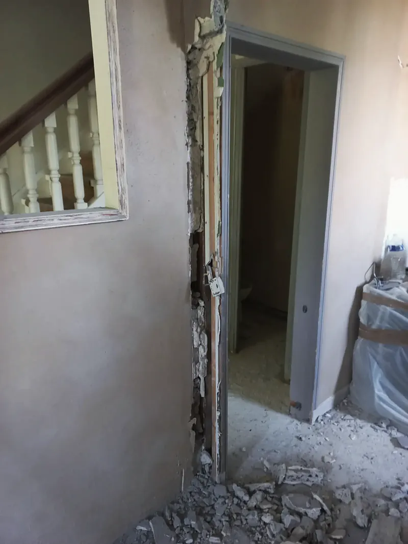 Mur intérieur avec une ouverture encadrée donnant sur un escalier à balustres blancs, encadrement de porte en cours de dépose, gravats au sol.