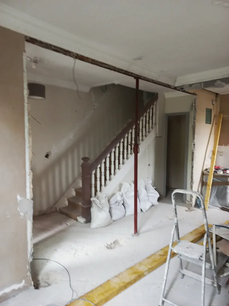 Large ouverture dans un mur, poutre au-dessus soutenue par un étai rouge, escalier à balustres au fond, sacs de gravats et escabeau.