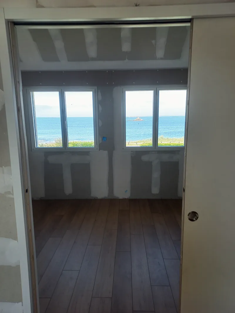 Chambre en travaux vue depuis une porte coulissante ouverte : deux fenêtres blanches donnant sur la mer, murs en plaques de plâtre aux joints enduits, sol en lames ton bois.