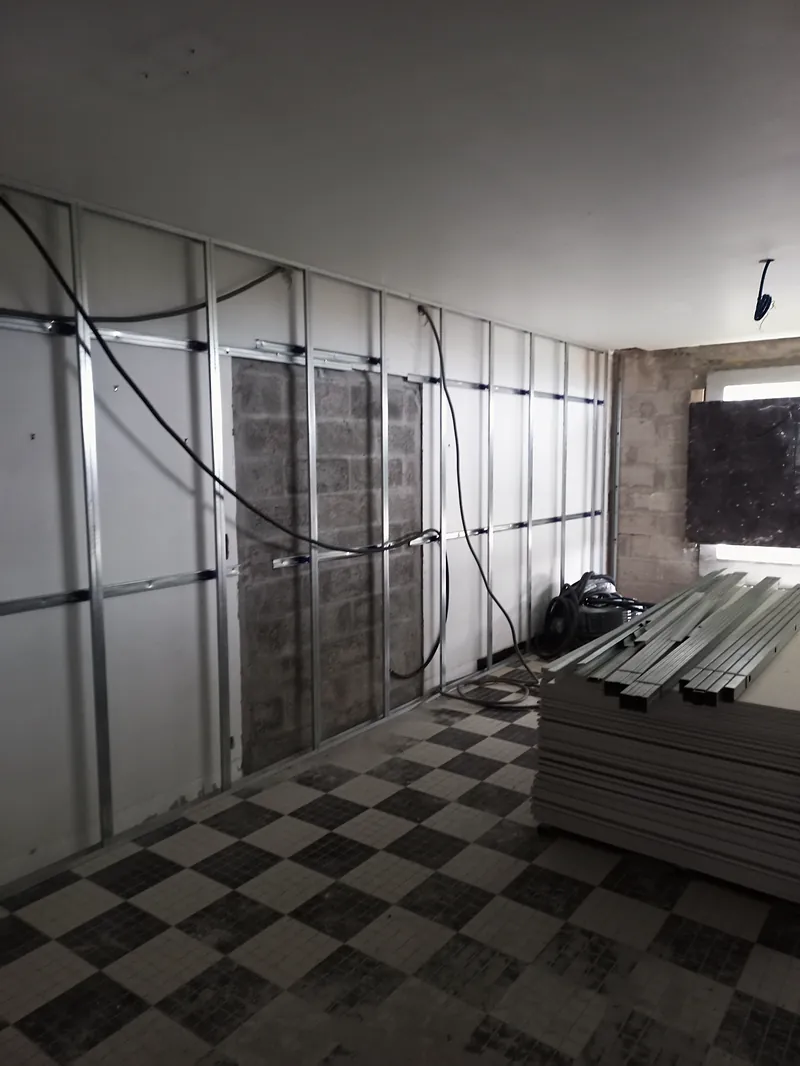 Pièce en travaux, montants métalliques fixés contre un mur en blocs béton, câbles électriques en attente, sol en mosaïque noire et blanche en damier, profilés et plaques empilés à droite.