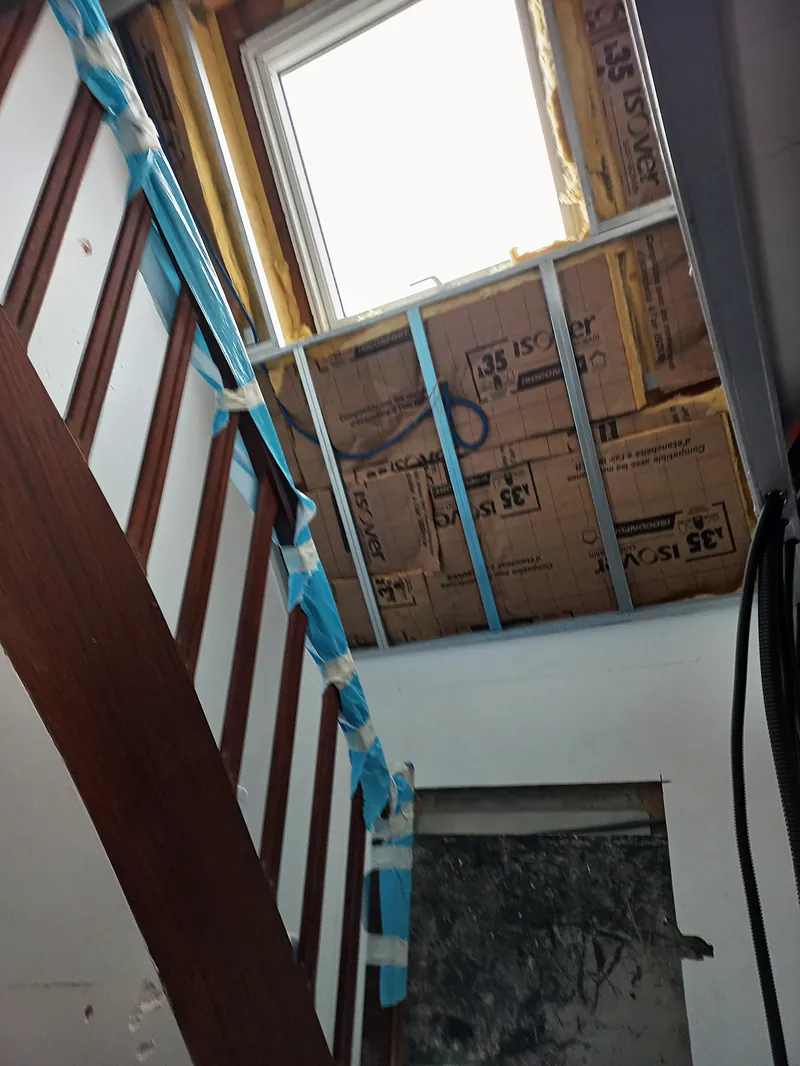 Cage d’escalier : garde-corps en bois protégé par un adhésif bleu, fenêtre de toit, panneaux d’isolant à parement kraft entre des montants métalliques.