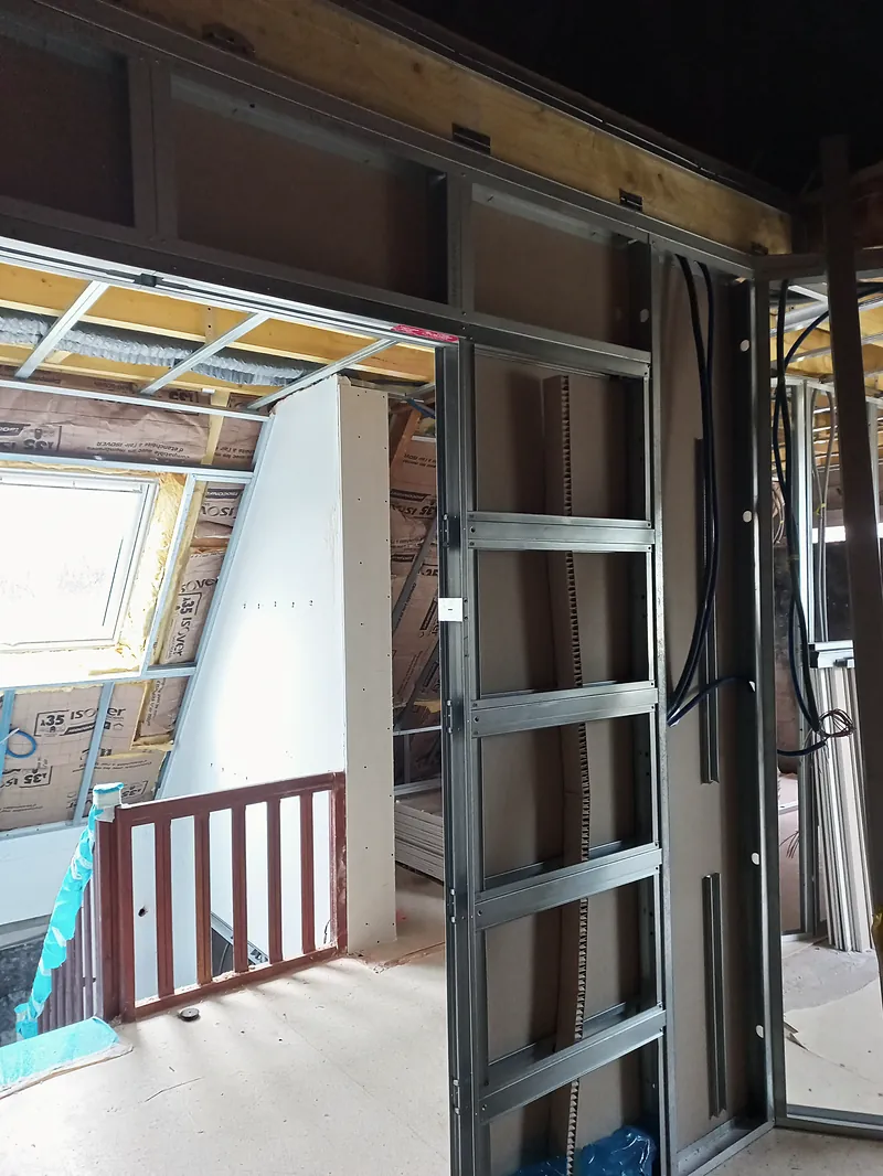 Châssis métallique de porte coulissante encastrée dans une cloison en plaques de plâtre, garde-corps en bois de l’escalier, fenêtre de toit et isolant au fond.