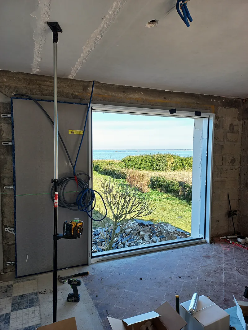Grande baie vitrée dans un mur en blocs béton, encadrée de plaques de plâtre, ouvrant sur une pelouse, une haie et la mer ; niveau laser sur une perche, outillage au sol.