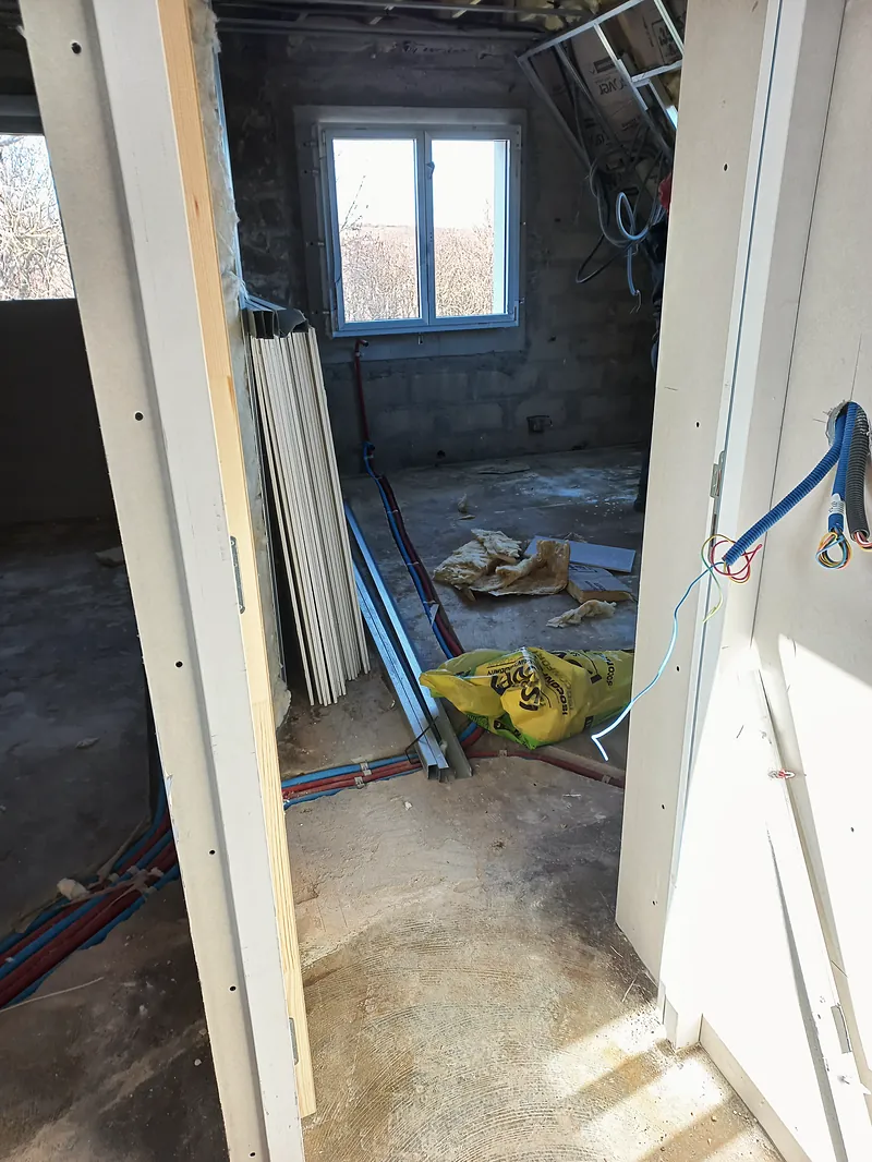 Pièce en travaux entre deux cloisons en plaques de plâtre, tubes rouges et bleus posés au sol, fenêtre sur des arbres au fond, profilés et sacs au sol.