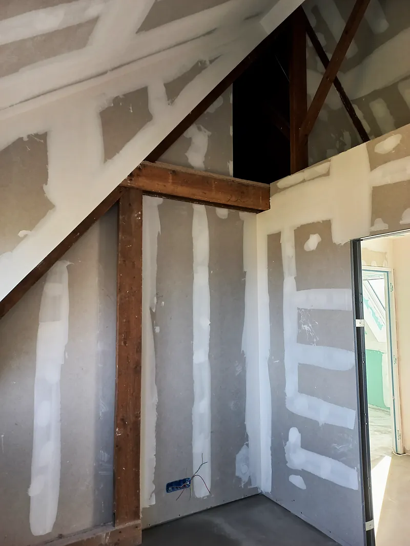 Chambre sous rampant aux plaques de plâtre jointoyées, poteau et poutre en bois brun laissés apparents, porte ouverte sur une pièce voisine.