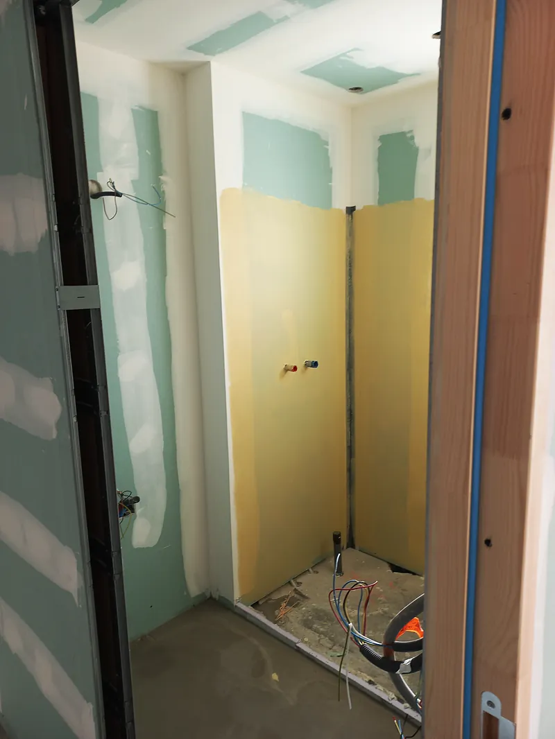 Emplacement de douche entre des cloisons en plaques vertes, membrane d’étanchéité jaune sur les murs, sorties d’eau en attente, sol brut.