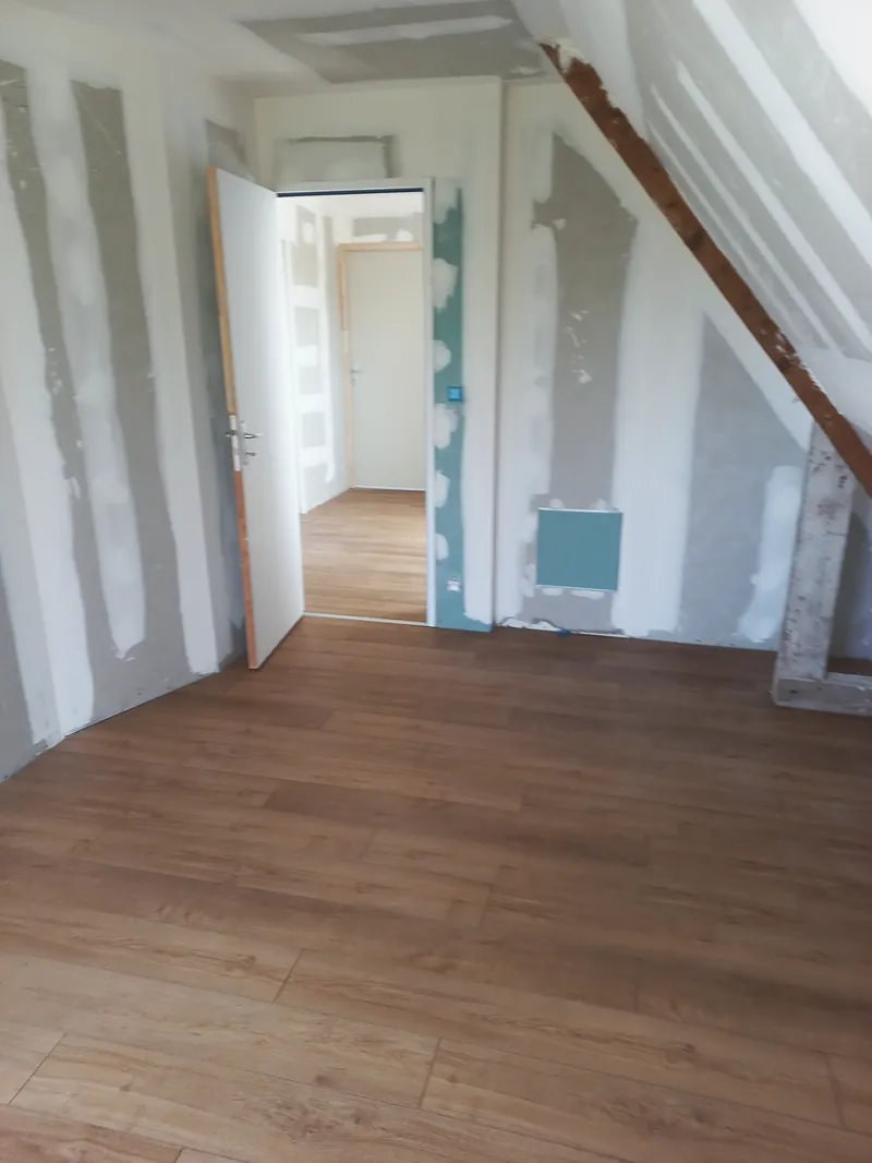 Chambre sous rampant au sol en lames ton bois, porte ouverte sur un couloir, plaques jointoyées, trappe de visite dans le mur.