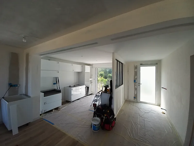 Cuisine en cours d’installation, meubles blancs sans plan de travail, verrière à profilés noirs le long d’un couloir menant à une porte d’entrée en verre dépoli, sol protégé.