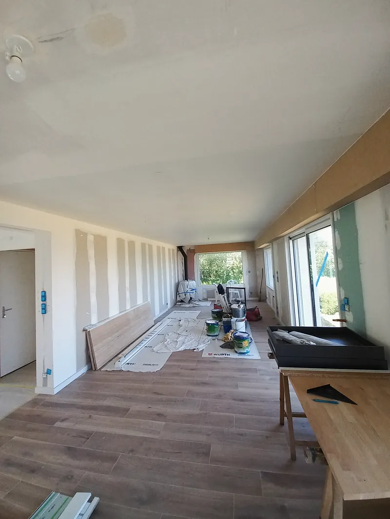 Longue pièce au sol en lames ton bois, murs aux joints enduits, baies vitrées à droite, pots de peinture et matériel sur une bâche, établi au premier plan.