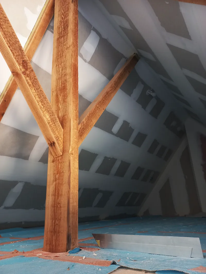 Poteau en bois et deux contrefiches sous des rampants doublés de plaques de plâtre aux joints enduits, sol couvert d’une membrane bleue.