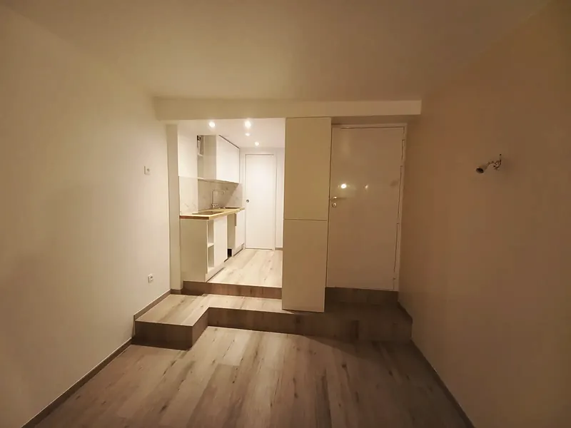 Appartement en longueur : estrade de deux marches menant à une cuisine blanche, porte de salle d’eau, sol stratifié clair.