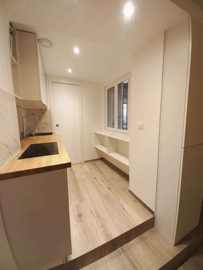 Cuisine étroite sur estrade, plan de travail bois et rangements blancs ouverts sous la fenêtre.