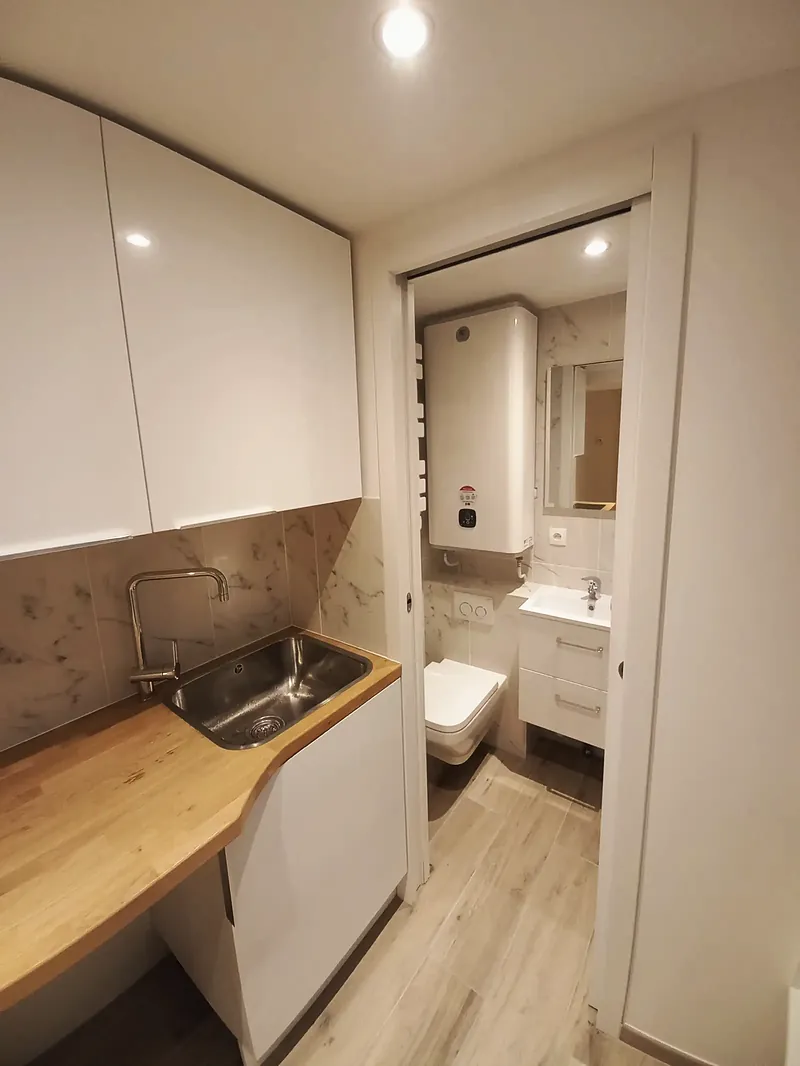 Évier de cuisine au premier plan, porte coulissante ouverte sur une salle d’eau avec WC suspendu et chauffe-eau mural.