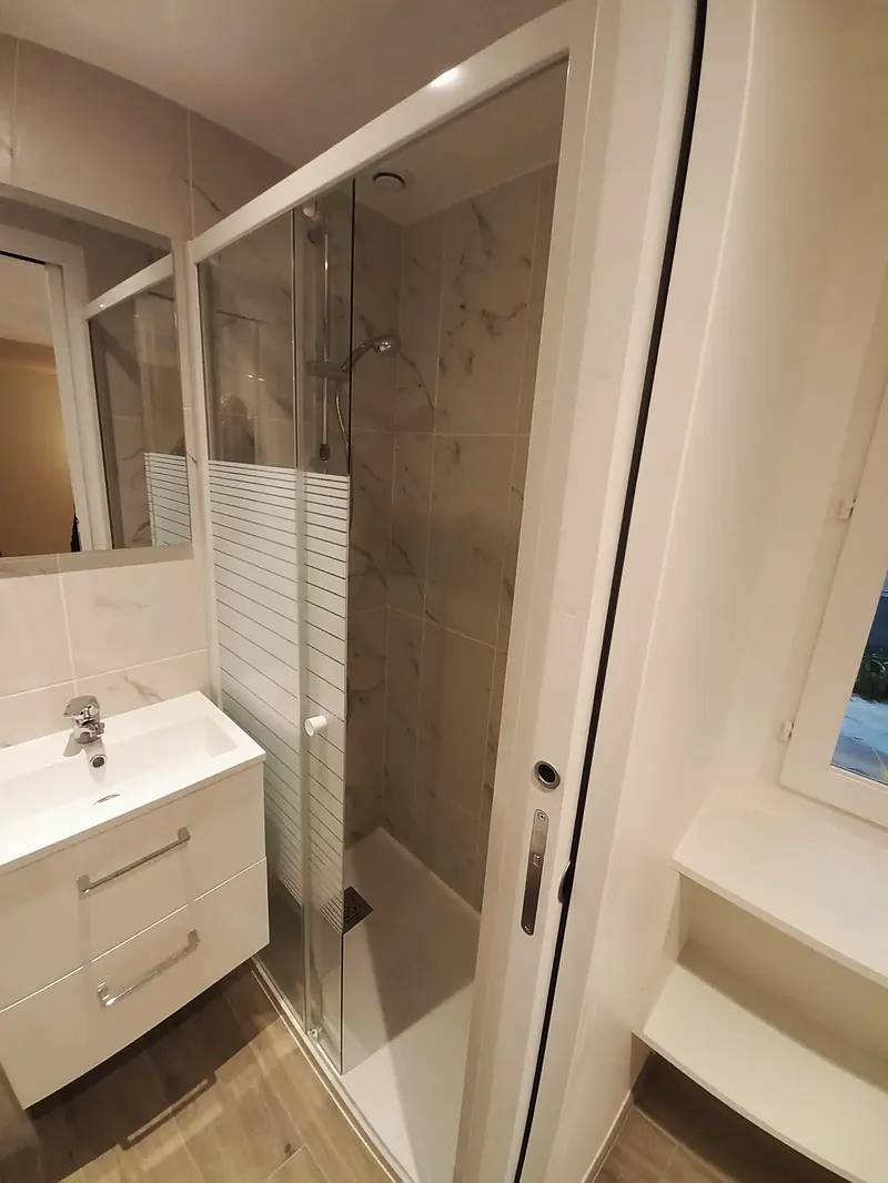 Salle d’eau avec douche à paroi vitrée, meuble vasque blanc et faïence effet marbre.