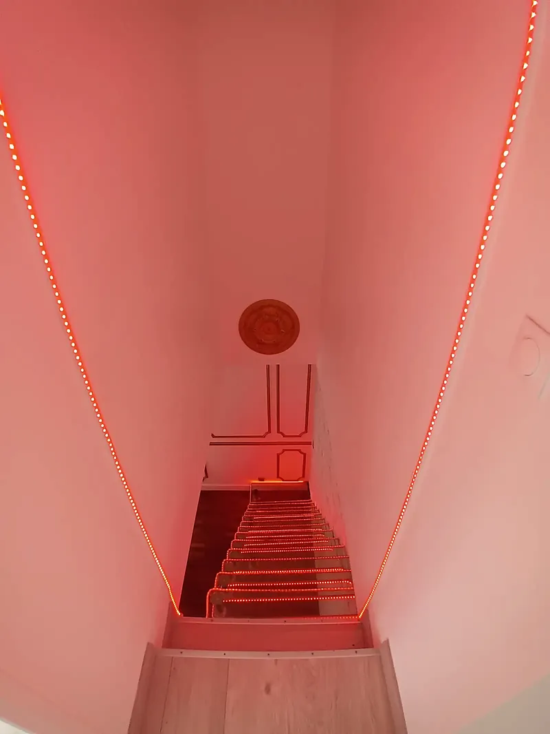 Escalier étroit vu d’en haut, murs éclairés par un ruban lumineux rouge.